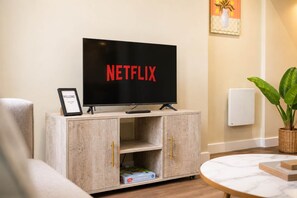 A television on a wooden cabinet with a 'NETFLIX' sign, a framed welcome sign, and a plant.