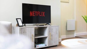 A modern living room with a flat-screen TV displaying 'NETFLIX' on the screen, a small cabinet with drawers, and a framed picture on the wall.