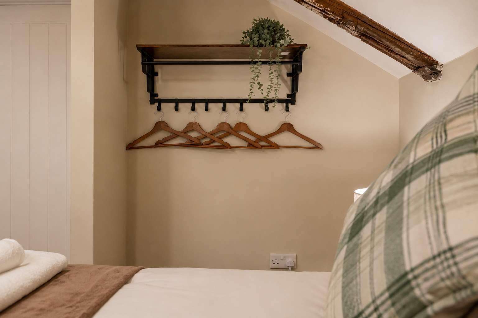 A bedroom with a bed, a wooden coat rack with clothes, a hanging plant, and a power outlet on the wall.