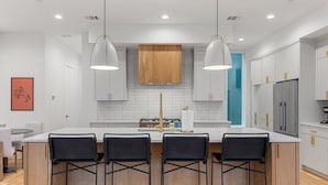 Una cocina moderna con armarios blancos, una isla central con taburetes de bar, iluminación colgante y un refrigerador de acero inoxidable.