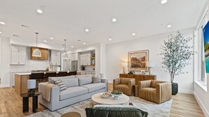 Una sala de estar moderna con un sofá, sillones y una mesa de centro. Se observa una cocina al fondo con armarios y una zona de comedor.