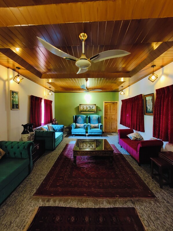 A spacious living room with a high ceiling, a ceiling fan, and a large window with red curtains. There are two sofas and a coffee table in the center.