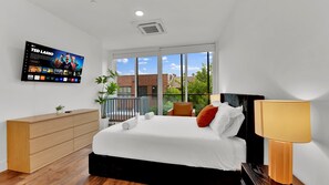 Une chambre d’hôtel moderne dotée d’un grand lit, d’un téléviseur fixé au mur, d’une commode en bois et d’un balcon offrant une vue sur les bâtiments et les arbres.