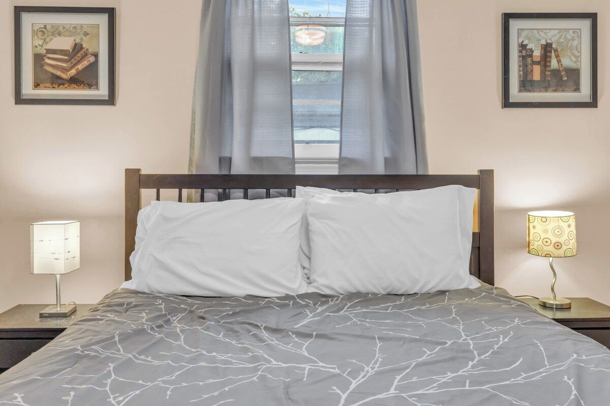 Una cama con una colcha gris estampada, dos almohadas blancas, un cabecero de madera, una mesita de noche con una lámpara y dos cuadros enmarcados en la pared.