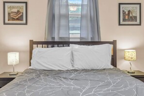A bed with a grey patterned bedspread, two white pillows, a wooden headboard, a bedside table with a lamp, and two framed pictures on the wall.