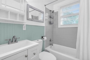 Een badkamer met een witte wastafel, toilet en bad. Er is een raam met zonwering, een spiegel boven de wastafel en een douche met een metalen rek.