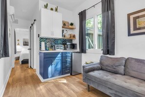 Un espacio habitable compacto con kitchenette, un sofá gris y una ventana con cortinas.