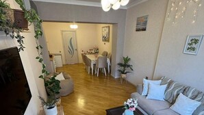Een woonkamer met een bank, salontafel en televisie. Er is een eethoek met een tafel en stoelen. De kamer heeft houten vloeren en decoratieve planten.