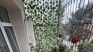 Een balkon met een houten tafel en twee stoelen, een witte airconditioning en potplanten.