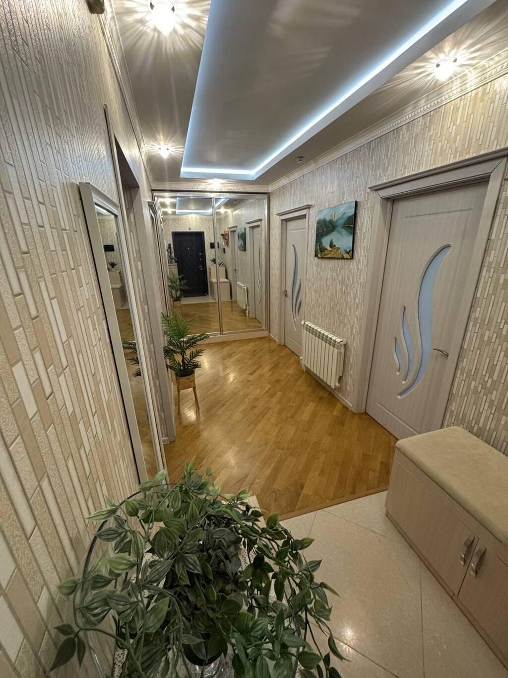 A hallway with wooden flooring, white walls, and a ceiling with recessed lighting. There are doors on the right, a bench on the left, and a large potted plant in the center.