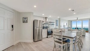 Una cocina moderna con electrodomésticos de acero inoxidable, un comedor con mobiliario de madera blanca y un amplio ventanal con vistas al océano.
