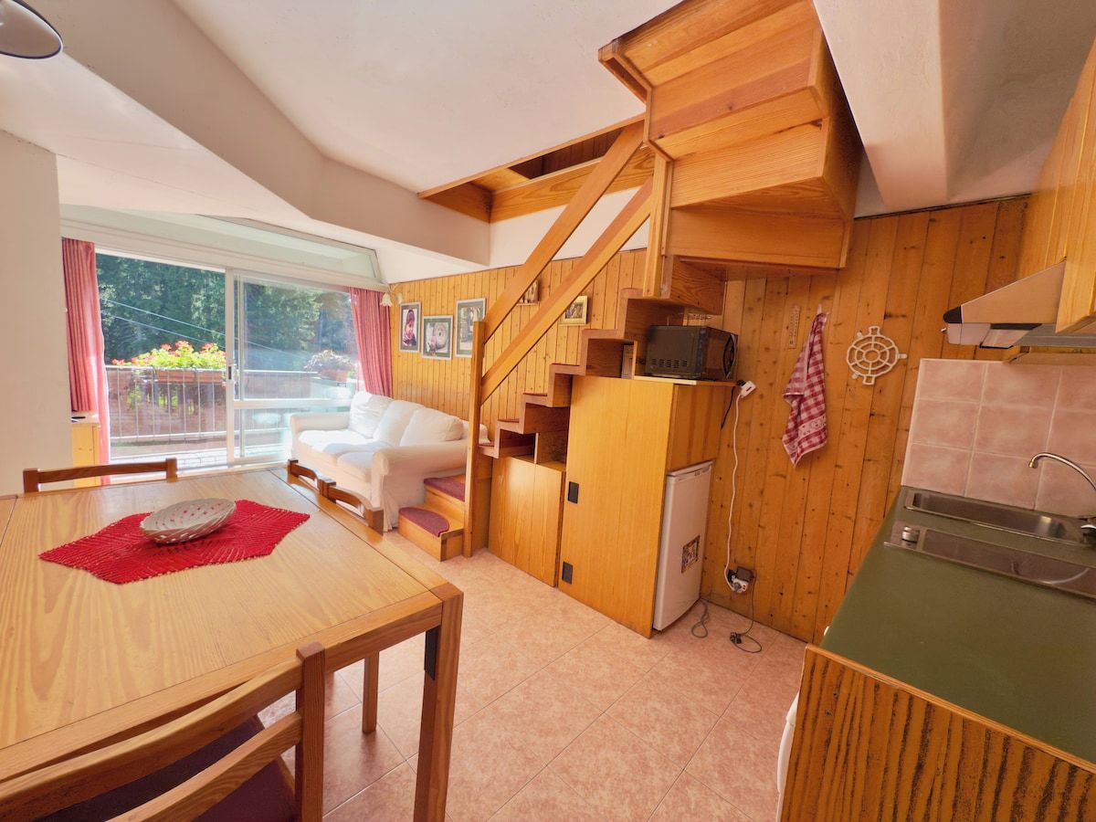 A compact kitchen with wooden cabinets, a dining area with a table and chairs, and a staircase leading to an upper level.
