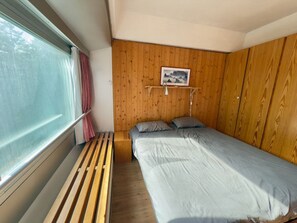 A bedroom with a bed, wooden panel walls, a window with curtains, and a bedside table.