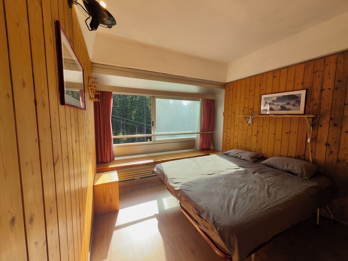 A bedroom with a large bed, wooden walls, a window with curtains, and a view of greenery outside.