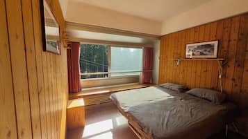 A bedroom with a large bed, wooden walls, a window with curtains, and a view of greenery outside.