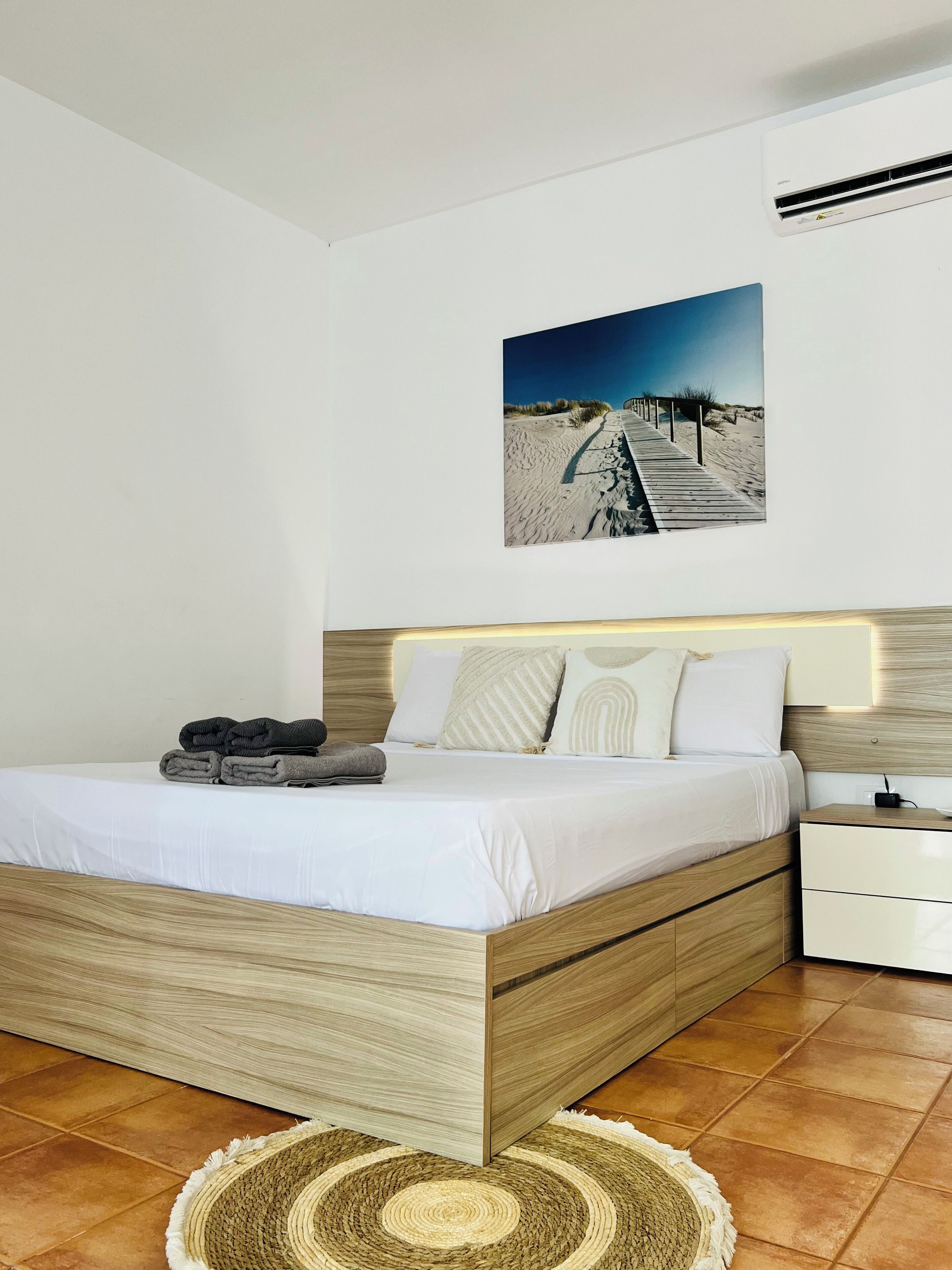 A modern bedroom with a wooden bed, white bedding, a headboard with a light fixture, a nightstand, and a beach-themed wall art.