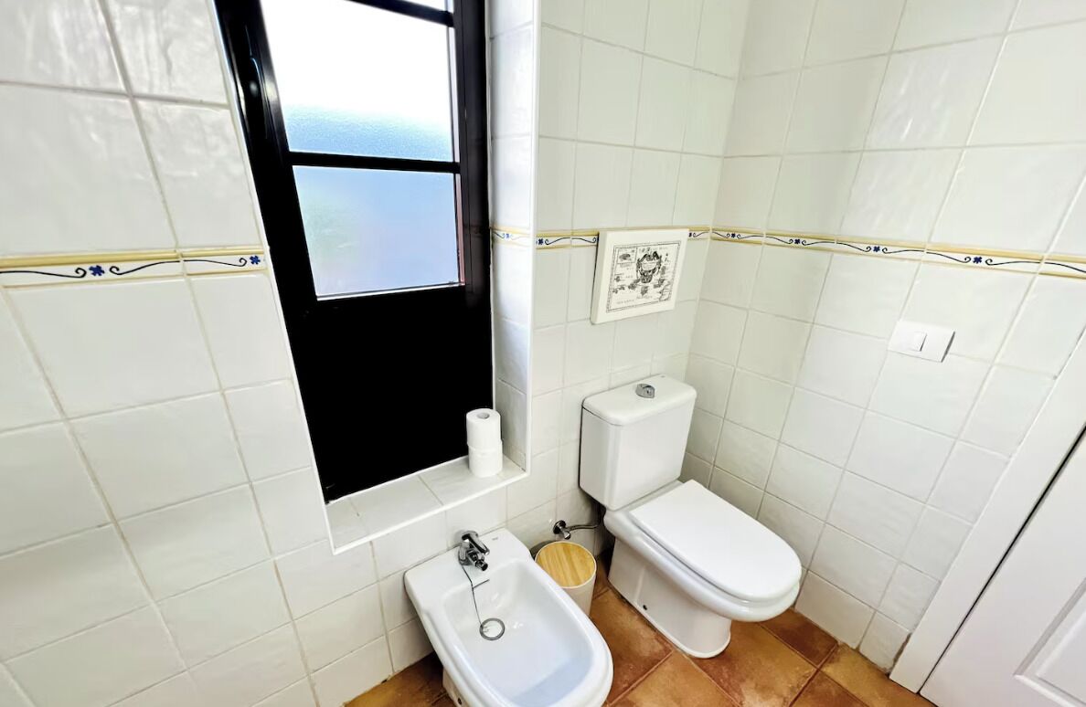 A small bathroom with white tiles, a toilet, a bidet, and a window.