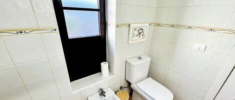 A small bathroom with white tiles, a toilet, a bidet, and a window.