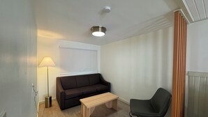 Ein kleiner, moderner Raum mit einer schwarzen Couch, einer Holzbank, einem grünen Sessel, einer Stehlampe und einer weißen Deckenleuchte.