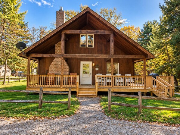 Front view of the cabin features a wide wooden porch with Adirondack chairs, a brick chimney, and rustic fence.