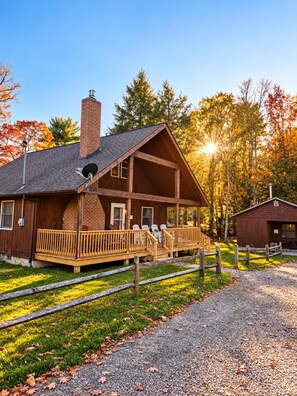A wooden lodge with a porch, a chimney, and a small adjacent building, surrounded by trees and a gravel path.