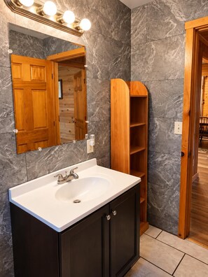 Bathroom features a white sink with dark wood vanity, marble-patterned gray tile walls, and a wooden shelving unit.