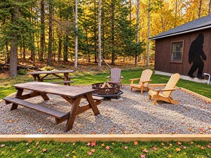 Outdoor fire pit area with three Adirondack chairs, two wooden picnic tables, and gravel ground cover.