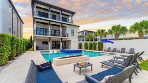 A modern outdoor pool area with lounge chairs, a fire pit, and a view of a multi-story building. - Walk to Beach + Pool, Hot Tub & Game Room 30A Ohana Grande by Avantstay (Santa Rosa Beach)