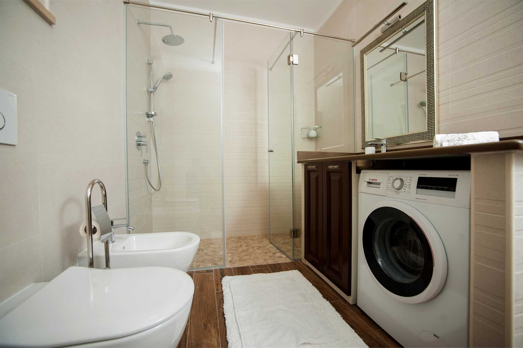 A modern bathroom with a glass shower enclosure, a Bosch washing machine, a toilet, and a sink.