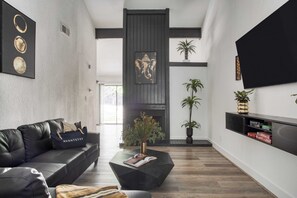 Una sala de estar moderna con un sofá de cuero negro, una mesa de centro negra y una chimenea. Hay plantas en macetas y un televisor de pantalla plana montado en la pared.