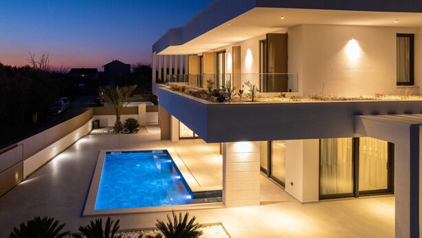 Una casa moderna de dos pisos amb piscina, grans finestres i exterior ben il·luminat al capvespre.