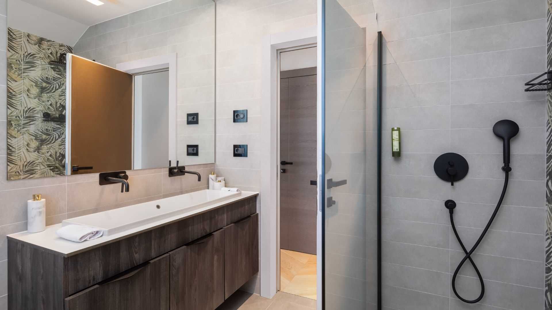 A modern bathroom with a large mirror, a dark wood vanity, a white sink, and a glass-enclosed shower area with a black hose.