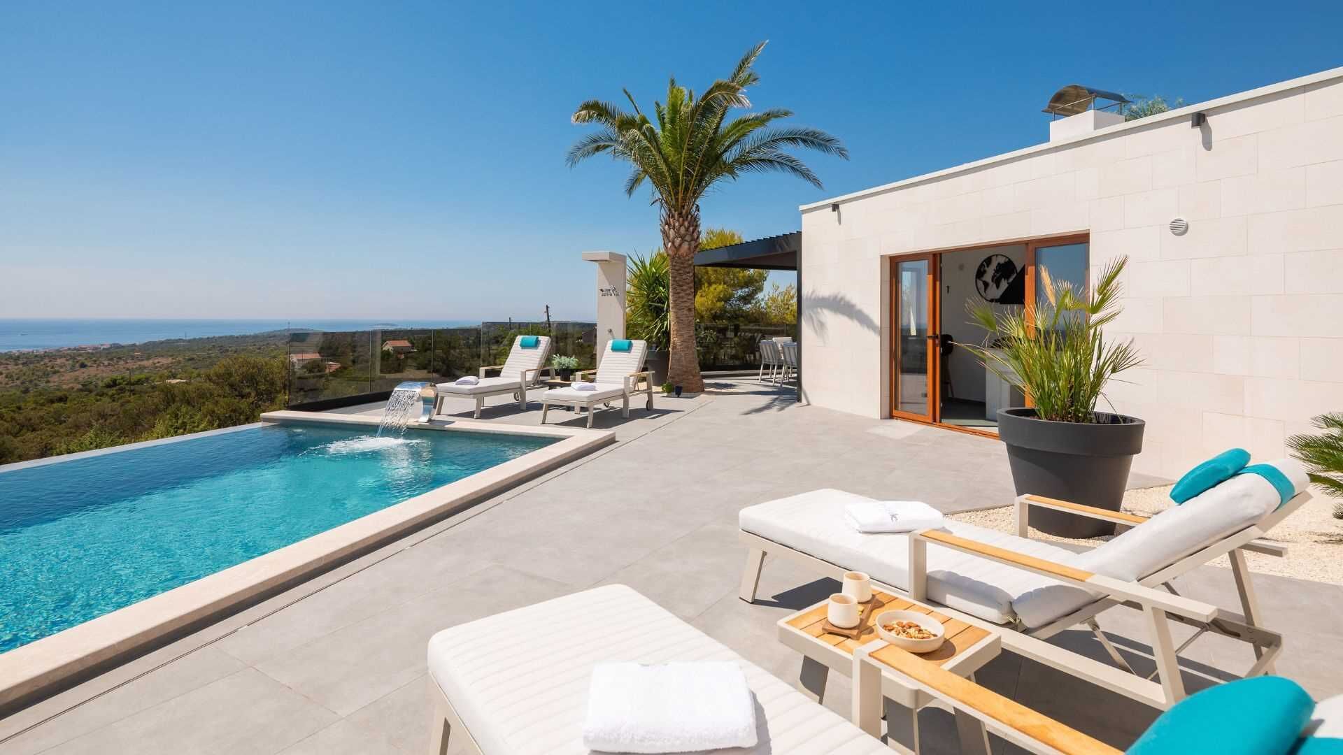 A modern outdoor pool area with lounge chairs, a small table with a bowl of nuts, and a view of the sea.