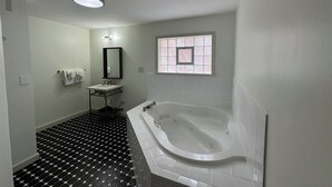 A bathroom with a black and white tiled floor, a white bathtub, a sink with a mirror, and a window with a grid pattern. - Non-Smoking King With Jacuzzi - NKJ (Manitou Springs)