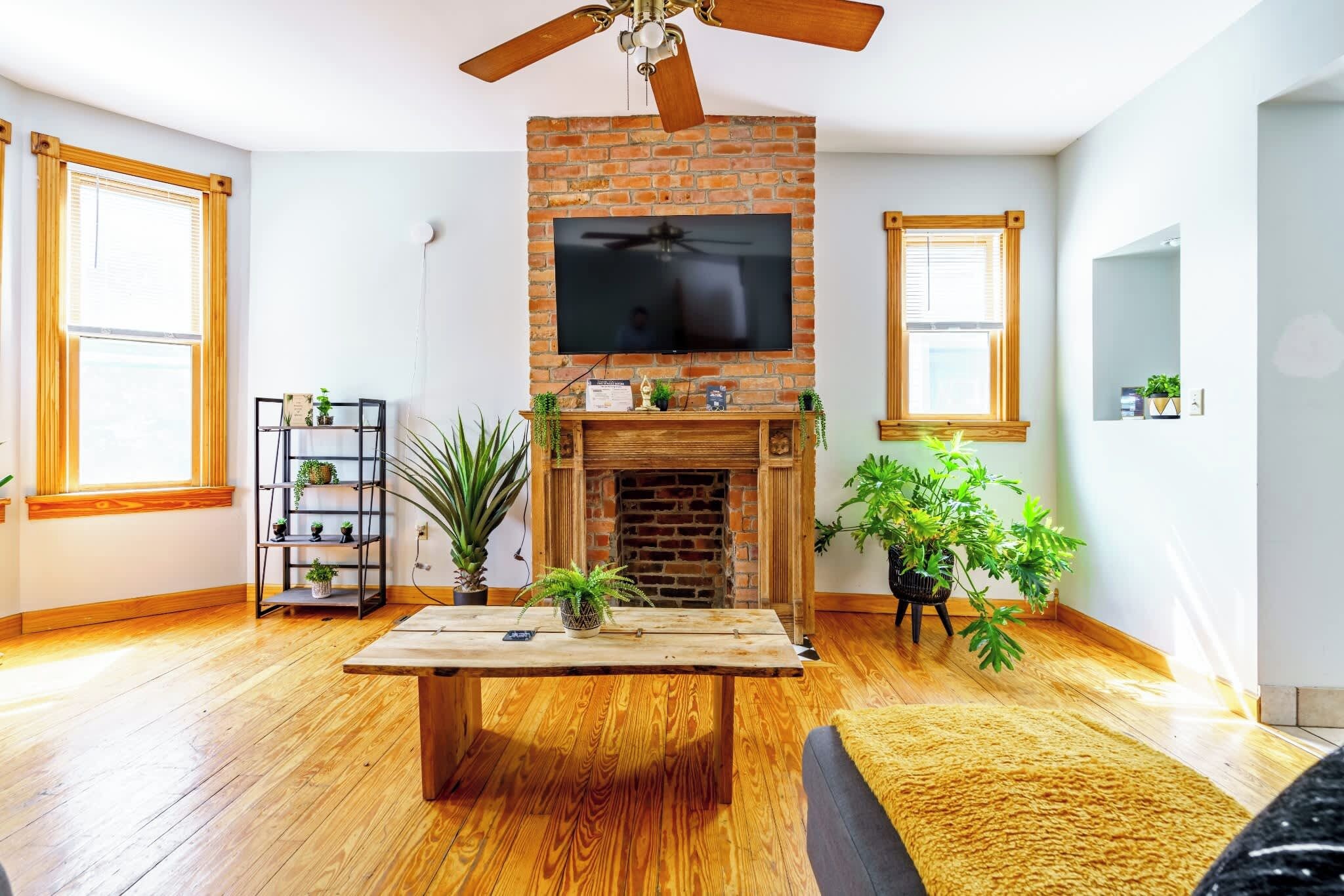 A living room with a brick fireplace, wooden flooring, a ceiling fan, and a flat-screen TV mounted above the fireplace.