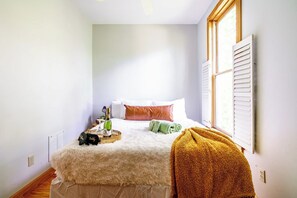 Un dormitorio con cama, una ventana con persianas, una mesita de noche con una botella, vasos y una bandeja.