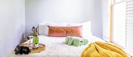 Une chambre d’hôtel avec un lit, des oreillers, une bouteille de champagne, des verres, un panier et une couverture.