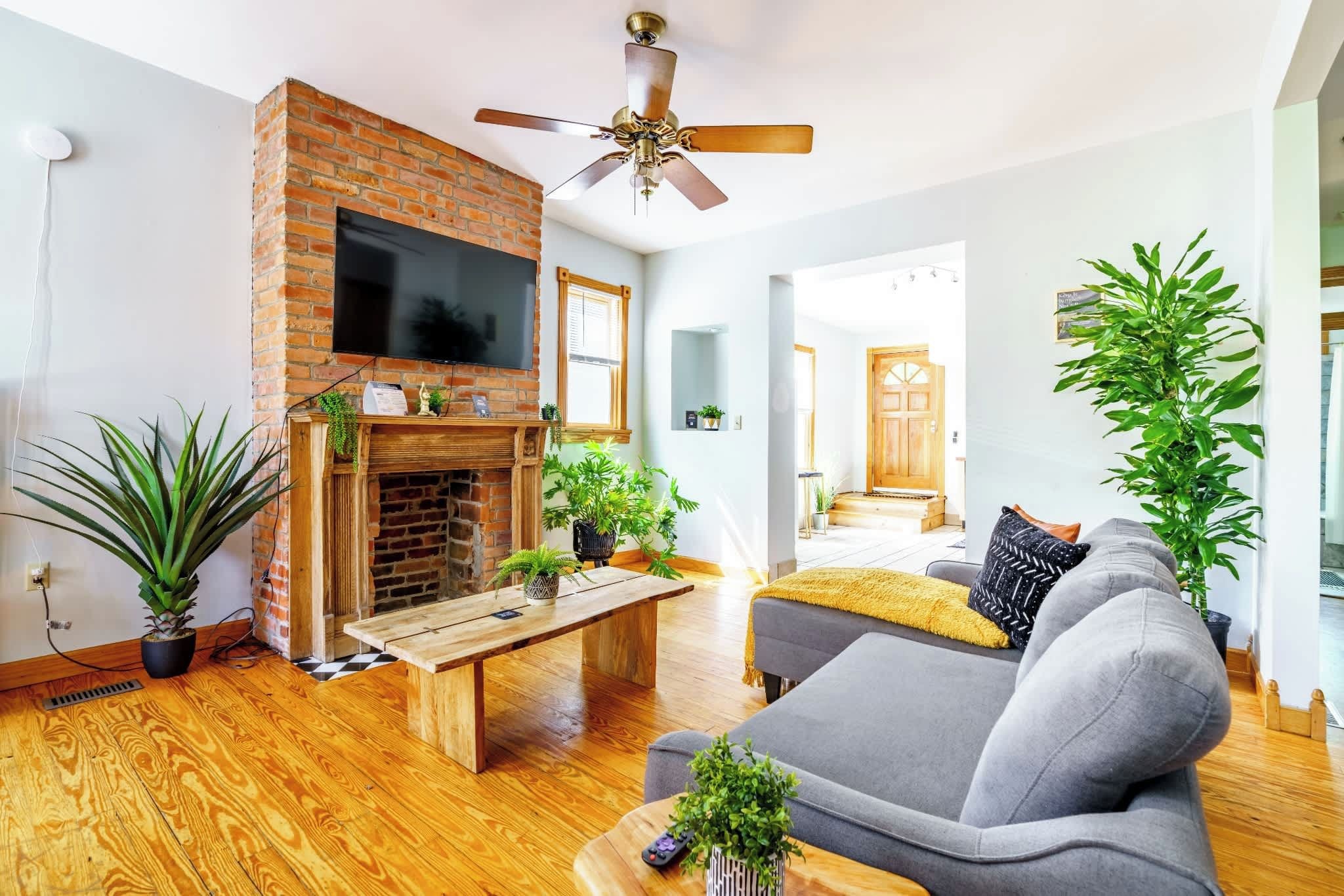 Un salon avec une cheminée en briques, un ventilateur de plafond, un canapé gris et des plantes en pot.