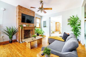 A living room with a brick fireplace, a ceiling fan, a grey sofa, and potted plants. - Short North & Downtown & OSU | Near High Street | Free Parking | 5,800+ Reviews (Columbus)