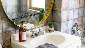 Ein Badezimmer mit einem weißen Waschbecken, einem spiegel mit goldener Rahmung, einem Seifenspender und einem kleinen Topf mit Pflanze.