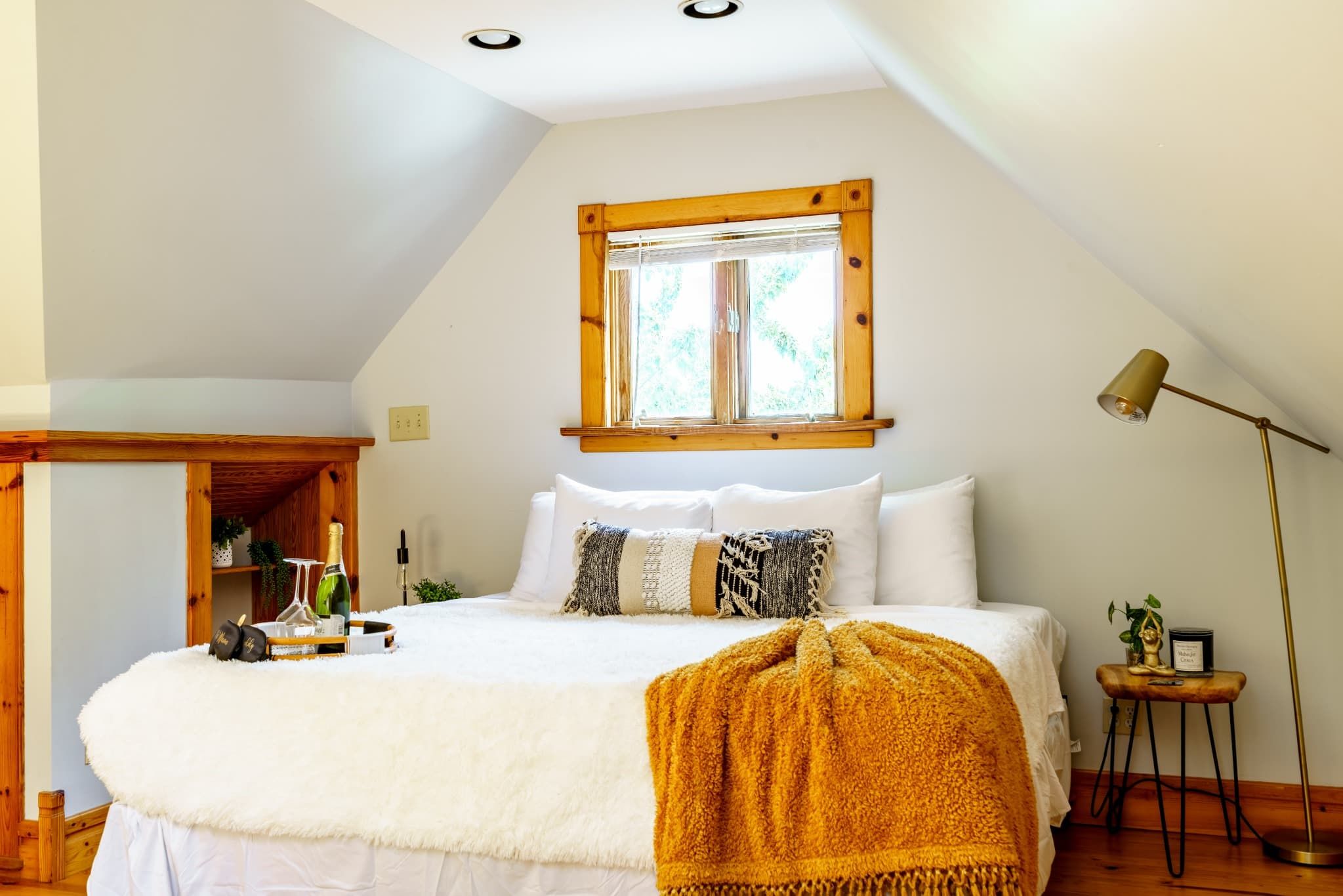 A bedroom with a large bed, a window with wooden framing, a bedside lamp, and a small table with a plant and a candle.
