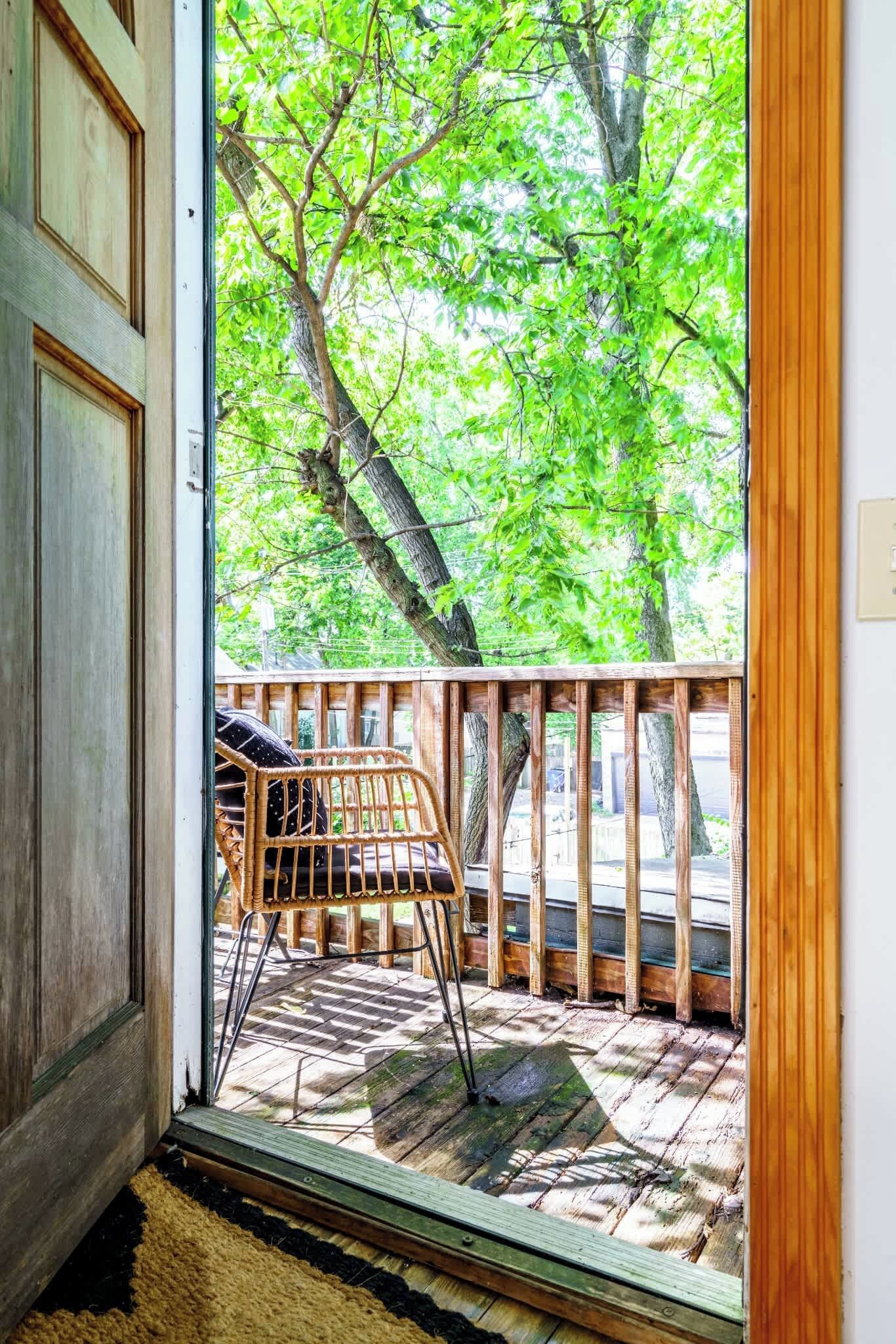 A wooden deck with a wicker chair and a view of green trees through a glass door.