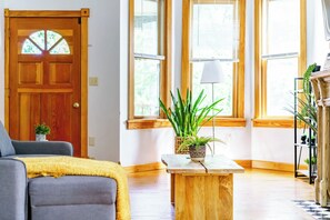 A living room with a wooden door, a sofa, a coffee table with a plant, and large windows with blinds. - Short North & Downtown & OSU | Near High Street | Free Parking | 5,800+ Reviews (Columbus)