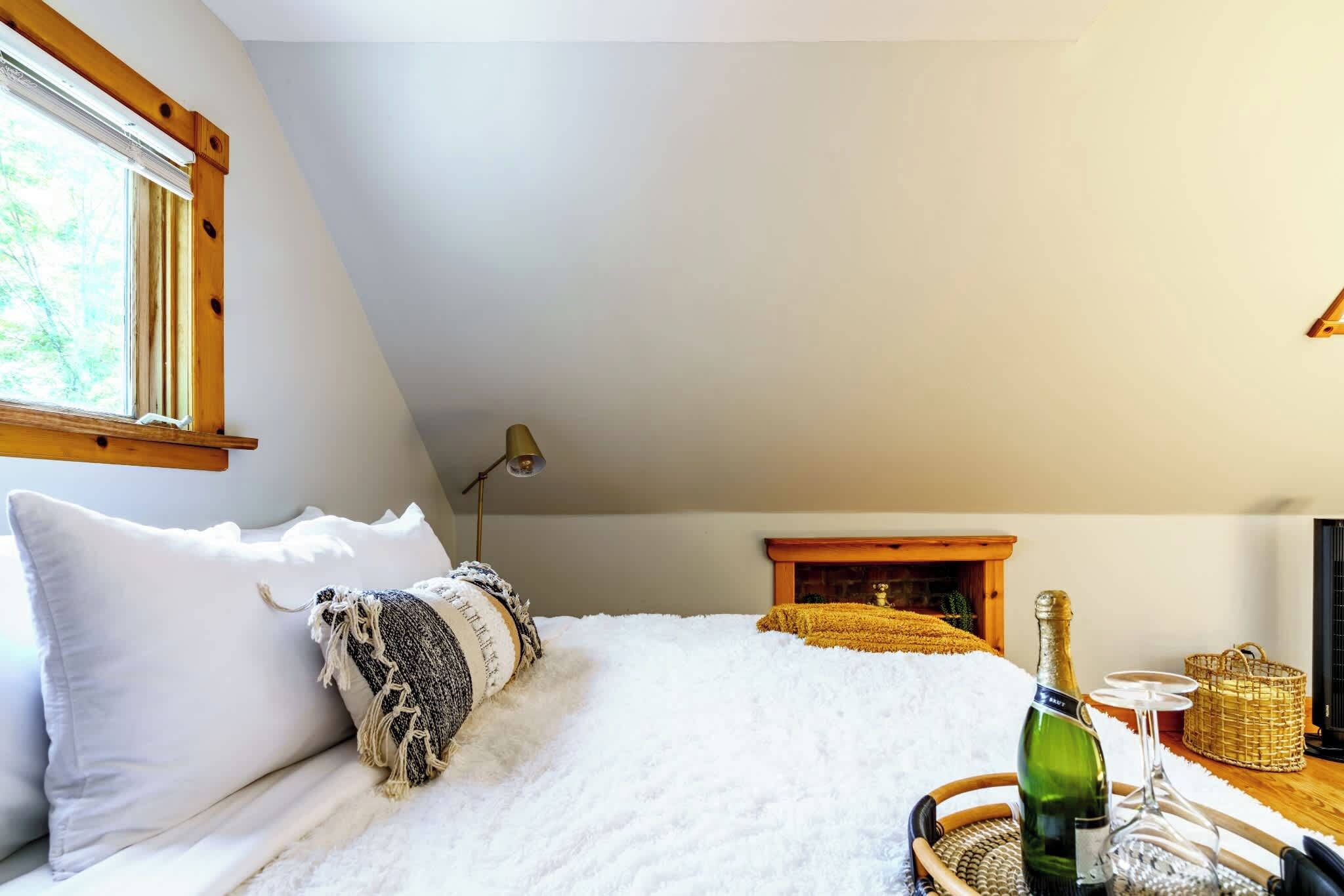 A bedroom with a bed, pillows, a lamp, a fireplace, and a bottle of champagne.