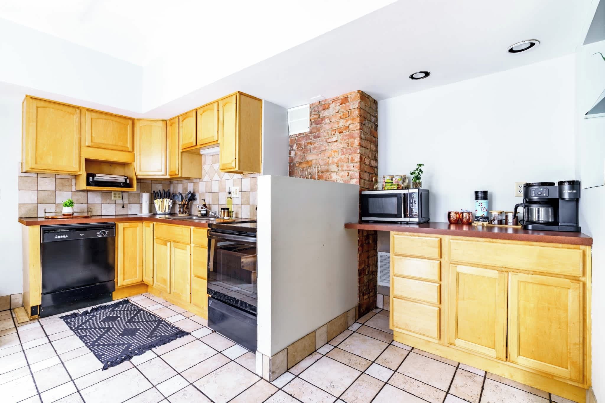 Una cocina compacta con armarios de madera, pared de ladrillo, microondas, cafetera y lavavajillas.