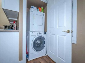 A compact laundry room with a stacked washer and dryer, a white door, and a small countertop with cleaning supplies.