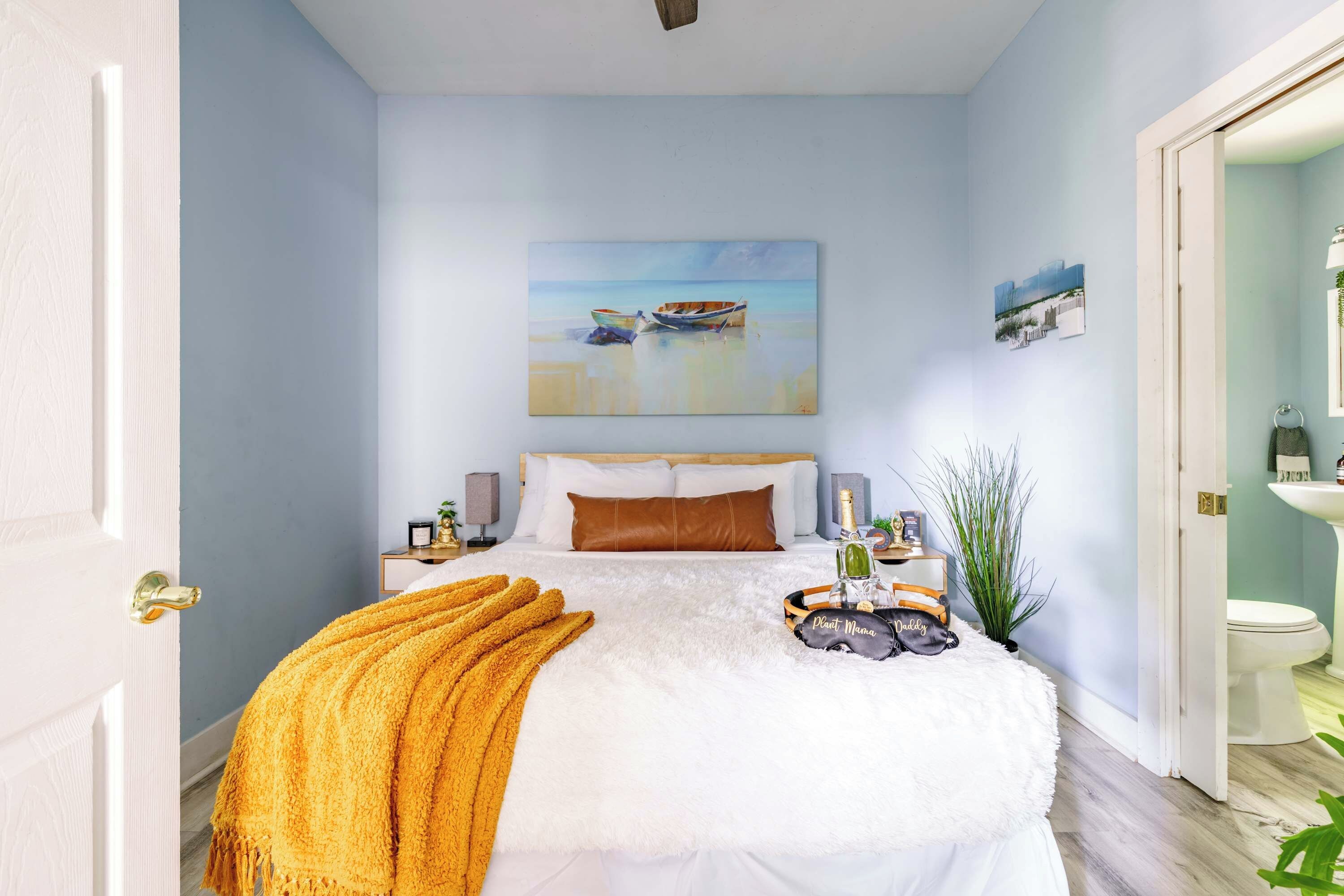 Un dormitorio con cama, un cuadro de una playa, una mesita de noche con lámpara y una mesilla.
