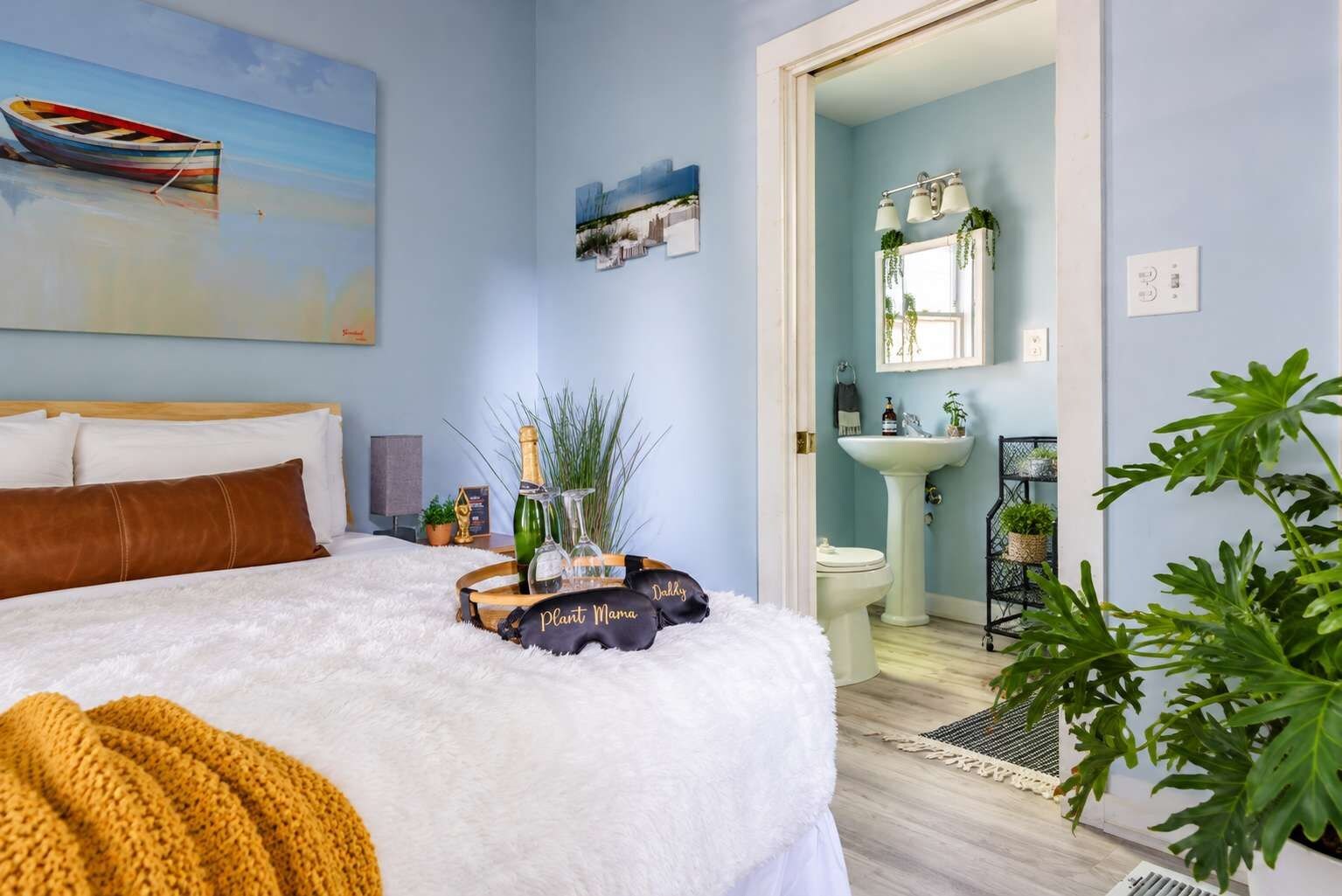 A bedroom with a bed, a bedside table with a bottle, glasses, and a plant, and a view of the bathroom.