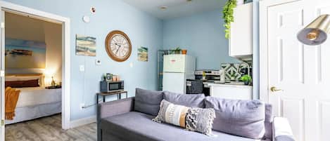 Een moderne woonkamer met een grijze bank, een klein tafeltje en een keuken met een koelkast en magnetron.