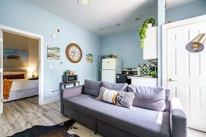 Ein modernes Wohnzimmer mit einem grauen Sofa, einem kleinen Tisch und einer Küchenzeile mit Kühlschrank und Mikrowelle.